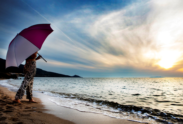 Schneider Schirme Easy in pink/white – with a removable ground spike and base plate, perfect for trips to the beach and open-air bath. A woman stands under the Schneider Easy parasol, gazing at the sea during sunset.