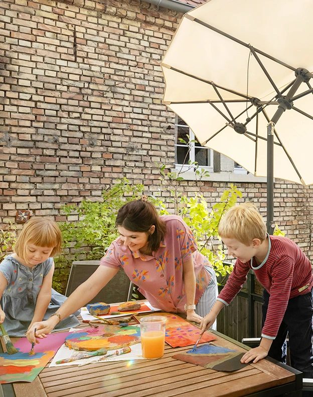 Schneider Schirme Salerno 350x150 cm in ecru in use - opens an closes easily in small areas. A woman is painting with watercolors at a table on the balcony with two children under the Schneider Schirme Salerno 350x150 cm in ecru. In the background, the brick wall of the house is visible.
