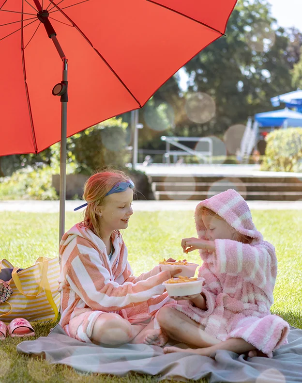 Schneider Schirme Locarno 220 cm Ø in red in use - available in multiple sizes, styles and colors. Two children in bathrobes sit on a mat in the shade of the Schneider Sonnenschirm Locarno, 220 cm Ø, in red, eating fries. In the background, the entrance to the swimming pool is visible.